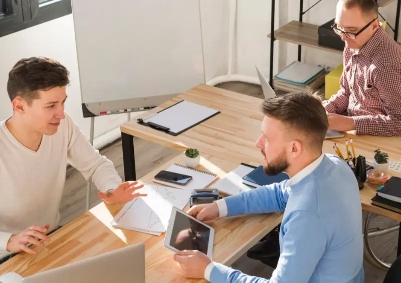 Team Collaboration Using a Tablet Three coworkers engaged in discussion at a shared desk, one demonstrating on a tablet for a business directory